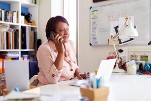 Mature Woman With Laptop Working In Home Office Using Mobile Phone
