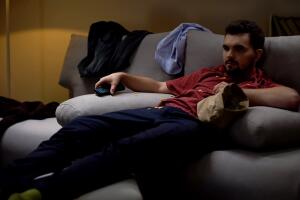 Messy man lying on sofa, watching tv, covered with chips crisps, untidiness