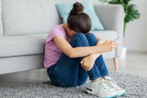 Depressed Indian teen girl with cellphone crying on floor at home, suffering from cyber bullying, being harassed online