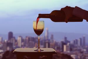 Female hand with bottle pours red wine into glasses on blurred San Francisco city background.