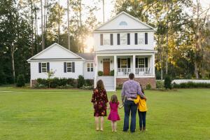 Family from behind standing outside front of new construction white siding farmhouse in the suburbs