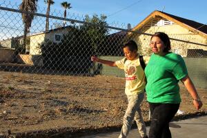 Idalia Ríos, una madre y vecina activista, camina a la escuela con su hijo Andrew, de 10 años, a través del vecindario Lacy de Santa Ana, en 2018. El lote baldío a sus espaldas, un área preocupante en una zona con contaminación por plomo, fue luego convertido por la ciudad en un parque, luego que activistas presionaran por más espacios al aire libre. (Yvette Cabrera / Center for Public Integrity) 