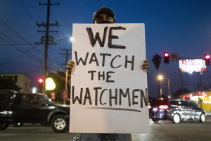 Un manifestante sostiene un cartel que dice 'We Watch the Watchmen'