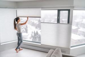 Woman opening home curtains in urban condo. Modern top down bottom up privacy cellular shades on apartment window keeping heat in winter with honeycomb blind curtain. Cordless pleated shades.