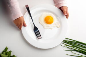 Children's hands hold a white plate with an egg, a fork and a knife.