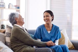 Consulting, caregiver and elderly woman laughing on sofa and holding hands in home living room. Support or healthcare, happy and female nurse talking or communication with senior citizen on a couch