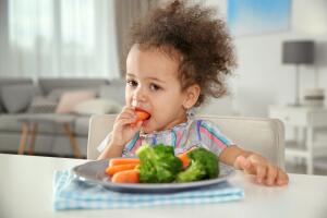 Cute African-American girl eating vegetables at table in living room