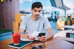 Skeptical male blogger amazed with incoming notification with strange message about interner changes in tariff plan on smartphone device sitting outdoors at wooden desktop with books and coffee to go