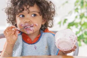 Toddler sitting in highchair and eating greek yogurt. Baby learning to eat and has yogurt on face and hair