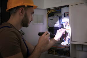 Young electrician with flashlight near switch box in dark room