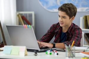 Schoolboy playing online game on laptop at home sitting table, teenage free time