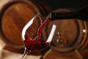 Pouring red wine from bottle into glass with wooden wine casks on background