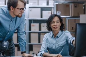 Male and Female Warehouse Inventory Managers Talking, Using Personal Computer and Checking Stock. Female Pointing at Screen. In the Background Rows of Shelves Full of Cardboard Box Packages.