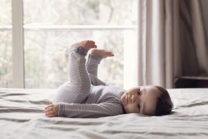 Side view of baby girl with brown hair wearing onesie lying on a bed, legs raised.